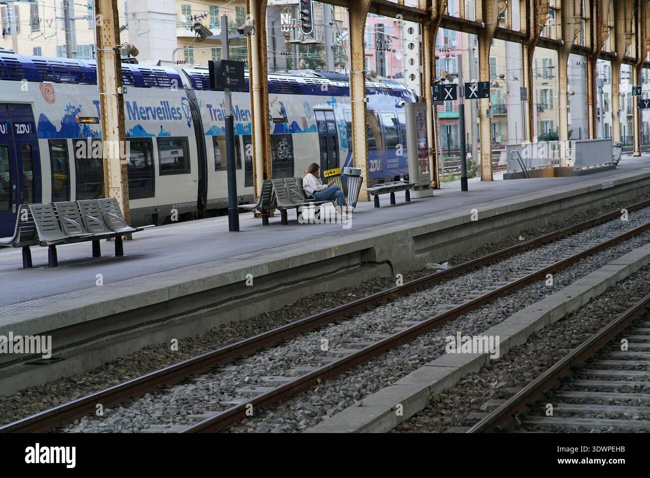 Ruhiger Moment am Gare de Nice Ville: Ein einsamer Reisender sitzt nachdenklich auf einer Bank unter goldenen Säulen, während ein blauer Zou 'Merveilles du Sud' Zug ruht Stockfoto