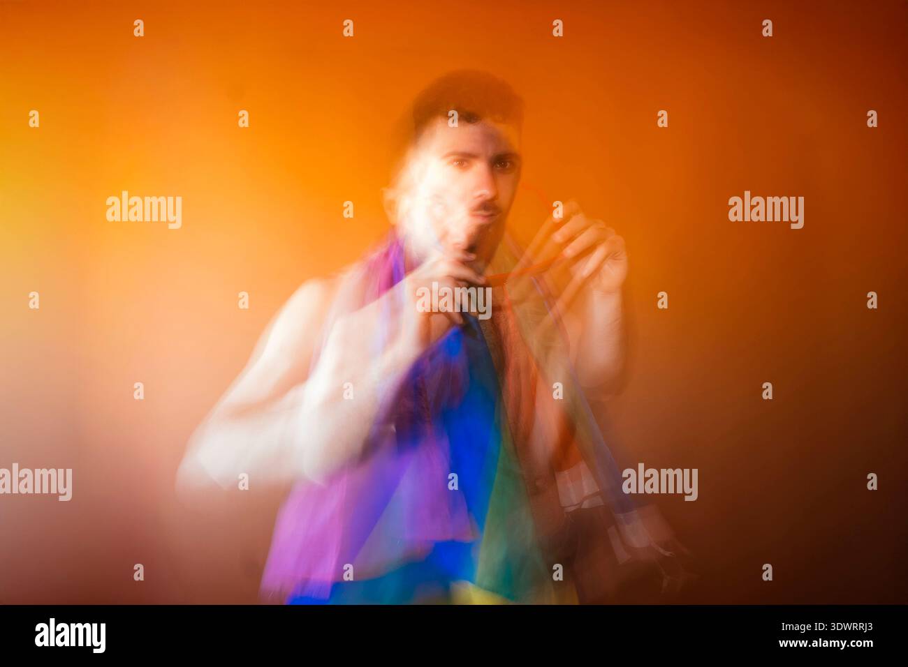 Junger Mann, der vor Bewegung unscharf steht und einen Regenbogenschal vor einem orangefarbenen Hintergrund hält Stockfoto