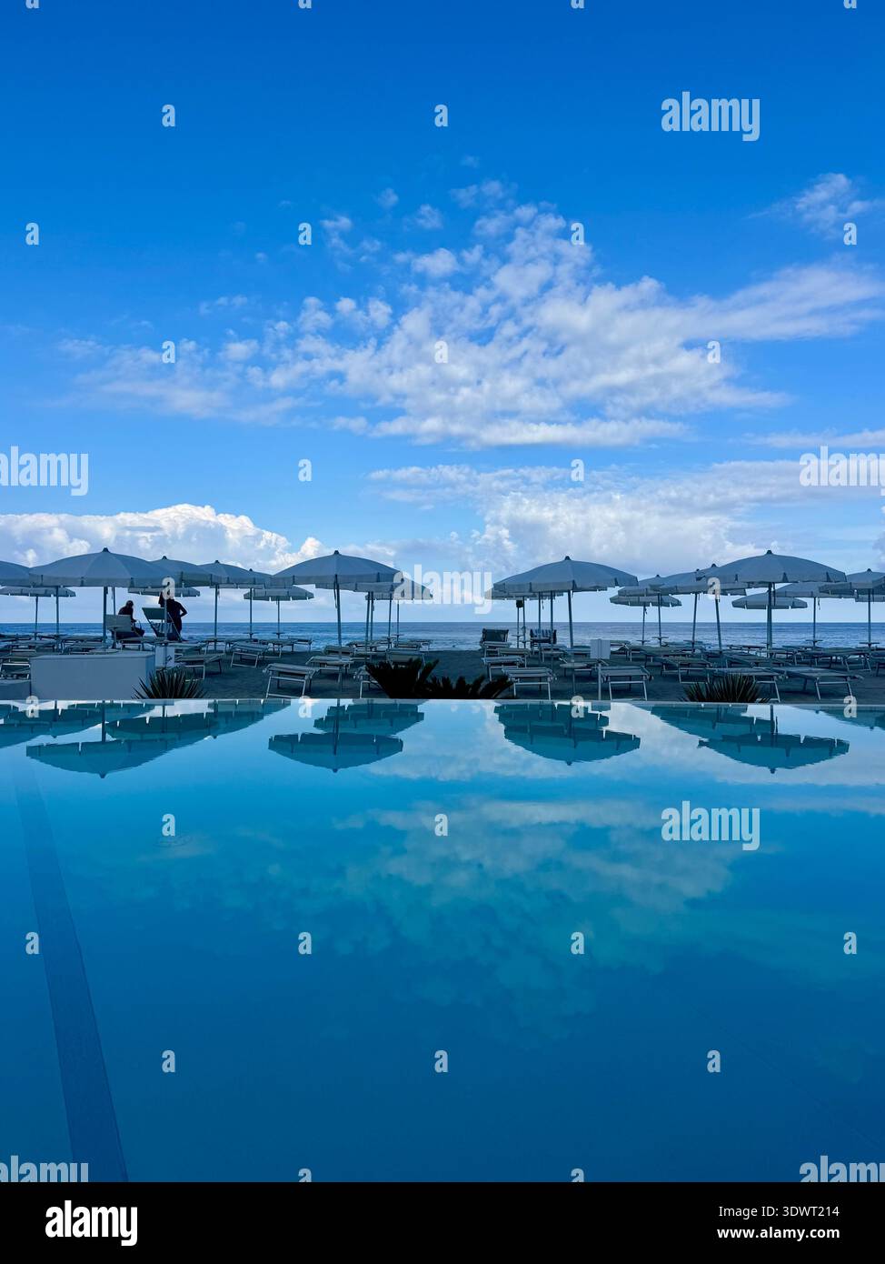 Sonnenschirme und Liegestühle am Meer spiegeln sich in einem ruhigen Swimmingpool unter blauem Himmel, ruhige Seebad Szene. - Smartphone-aufgenommenes Stockfoto