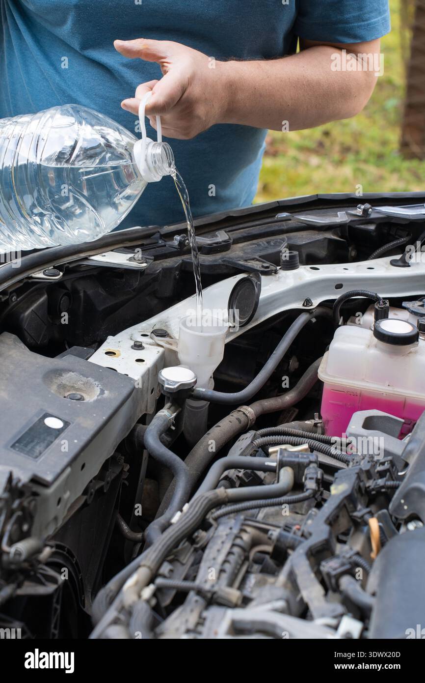 Eine Person gießt Scheibenwaschflüssigkeit aus einer Flasche in den Behälter eines Autos in einer Auffahrt. Die Sonne scheint und hebt die Autos hervor Stockfoto