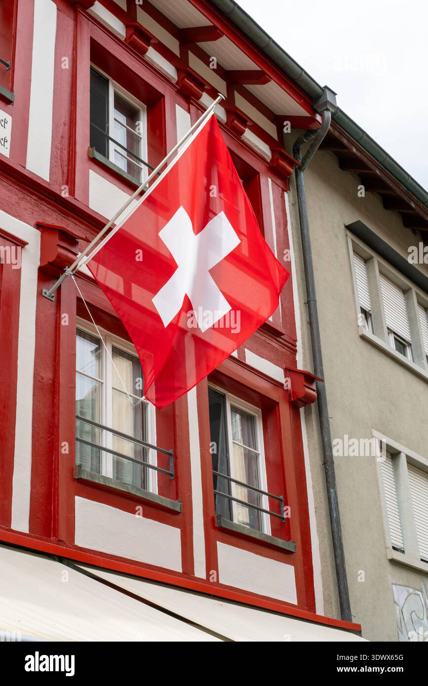 Eine große Schweizer Nationalflagge hängt an einem traditionellen Fachwerkgebäude. Die kräftigen Rot- und Weißtöne heben sich vom historischen Altstadtbogen ab Stockfoto