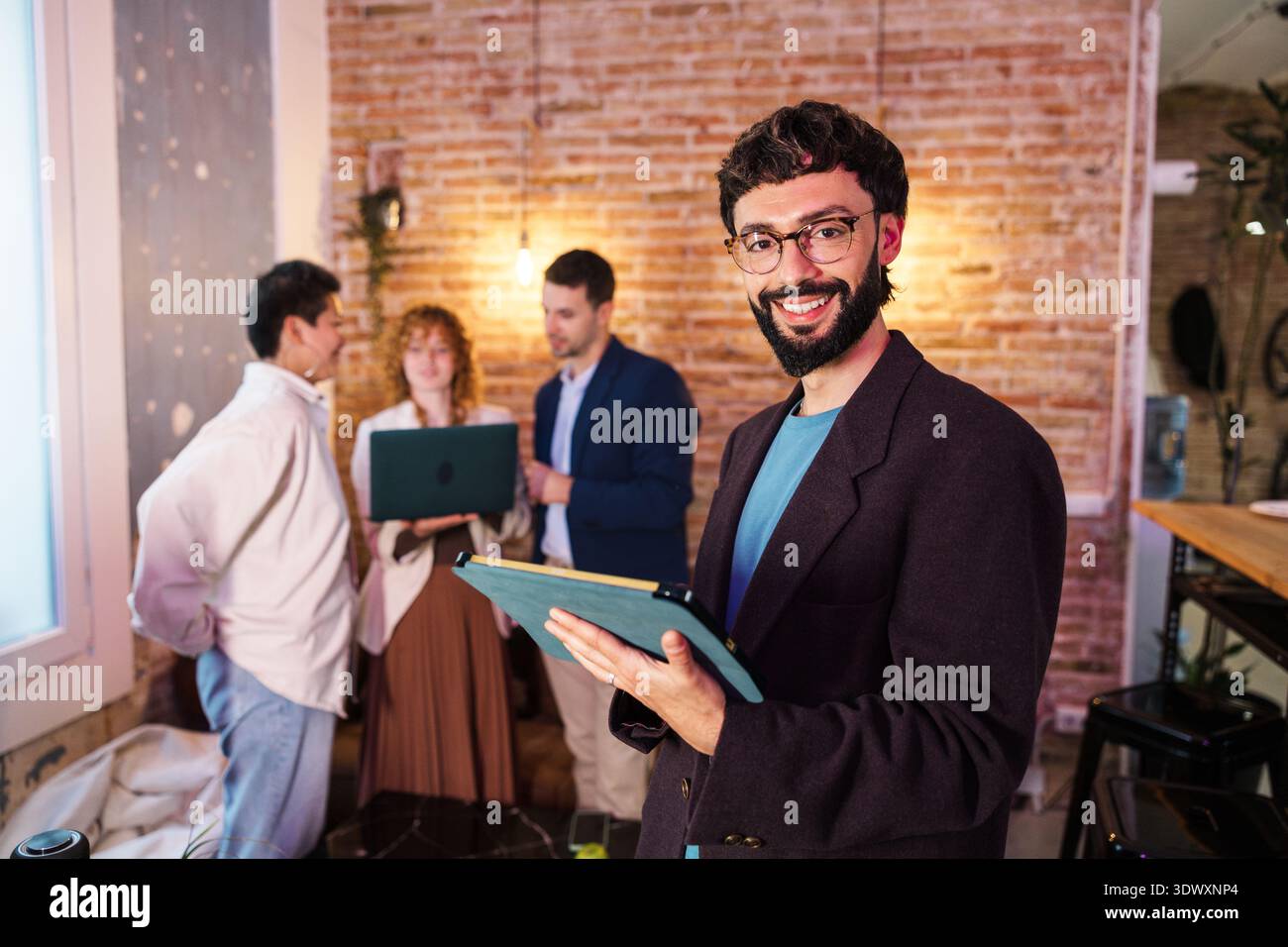 Lächelnder Geschäftsmann, der ein Tablet hält und Kollegen im Hintergrund arbeiten Stockfoto