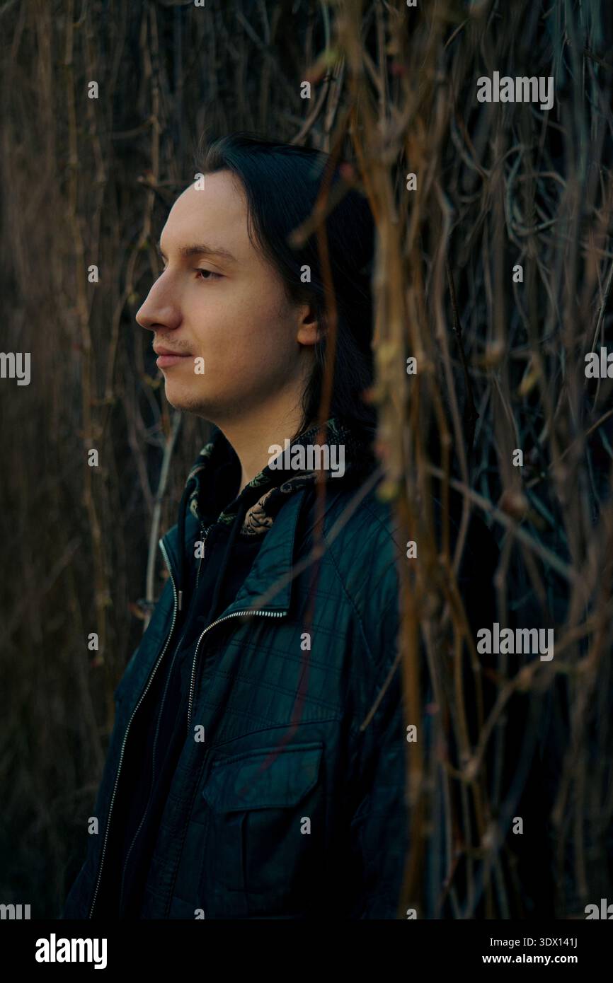 Seitenprofil-Porträt eines nachdenklichen jungen kaukasischen Mannes mit langen dunklen Haaren, in einer grau-blauen Jacke, stehend an einer trockenen Weinweinwand. Stockfoto