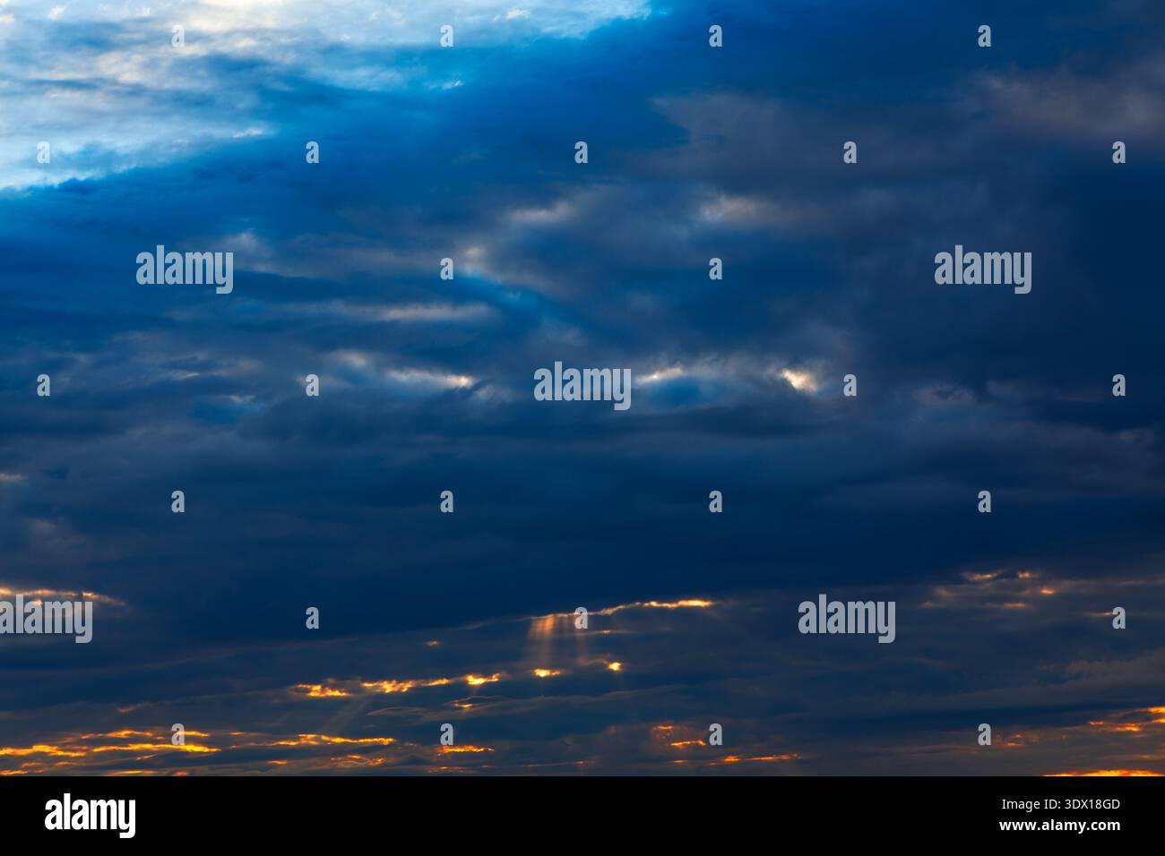 Der dramatische Himmel voller tiefblauer und grauer Wolken fängt das verblassende Licht des Sonnenuntergangs ein, mit goldenen Strahlen, die durch die Brüche hindurch ragen. Wolken rufen Ruhe hervor Stockfoto