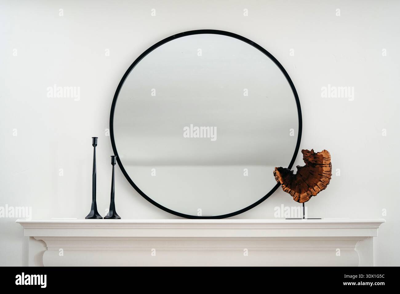 Minimalistischer Innenraum mit rundem, schwarz gerahmtem Spiegel über einem weißen Kaminregal mit dekorativen Objekten und neutralen Wandtönen. Stockfoto