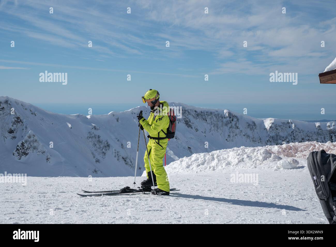 Russland, Krasnaja Polyana - 21. Februar 2026: Skifahrer im neongrünen Anzug justieren Stöcke auf verschneiten Bergrücken in 2200 Metern Höhe mit alpinen Gipfeln Stockfoto