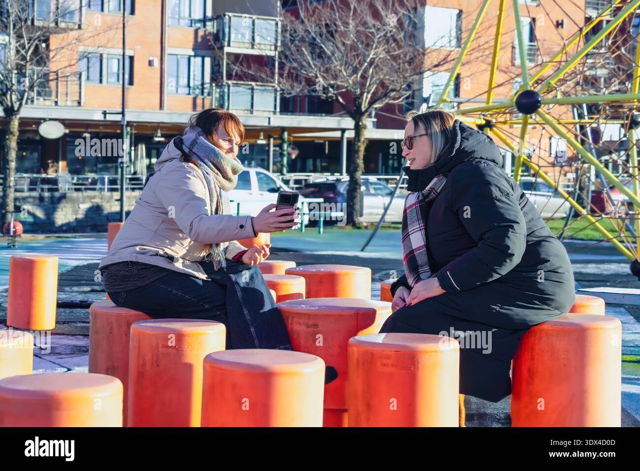 Zwei Frauen sitzen auf einem Spielplatz auf leuchtend orangefarbenen Hockern und teilen sich einen freudigen Moment, während sie ein Selfie machen, umgeben von einer lebhaften Outdoor-Atmosphäre Stockfoto