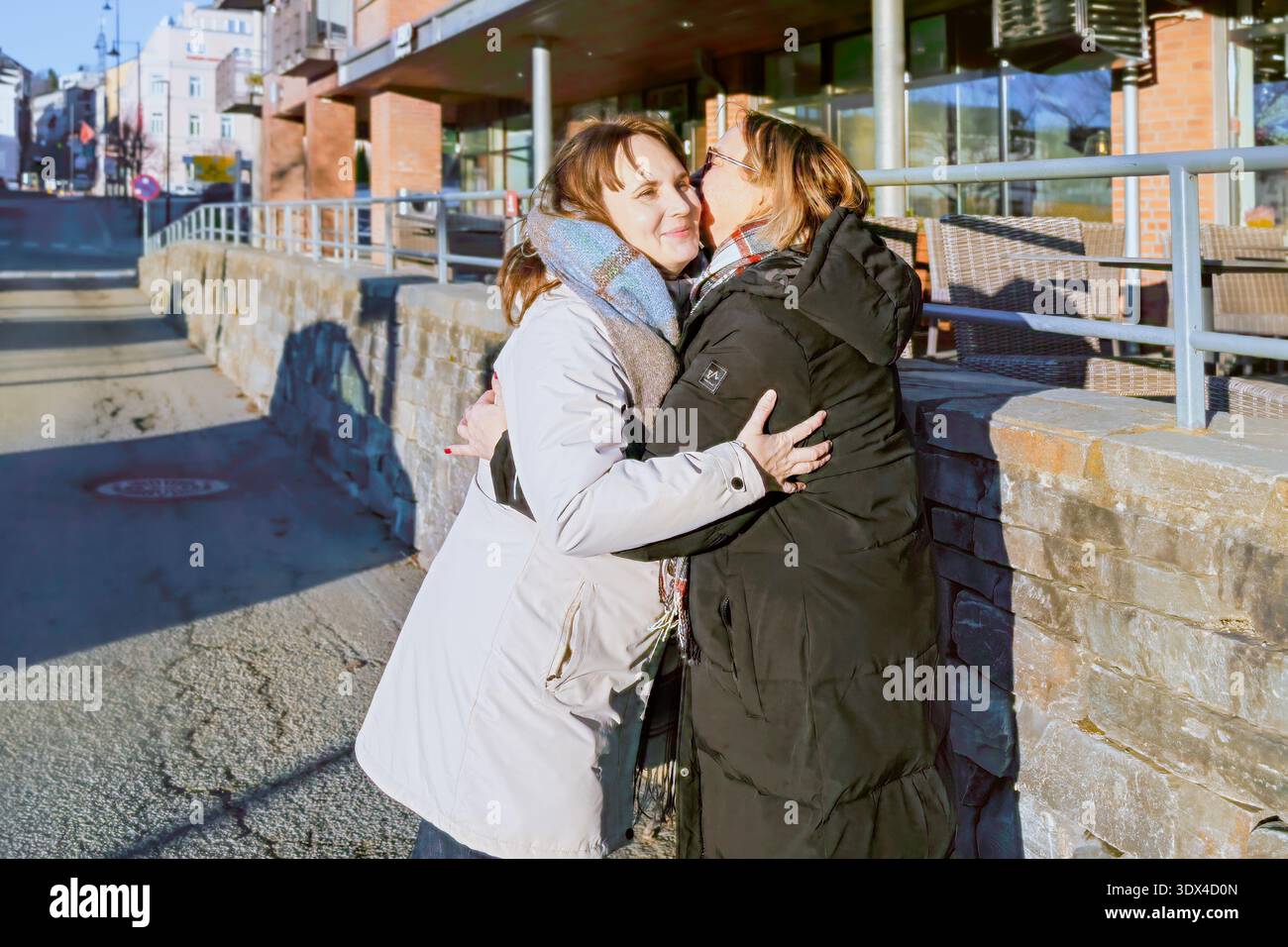 Zwei Frauen teilen sich eine herzliche Umarmung im Freien, tragen Wintermäntel, umgeben von einer urbanen Landschaft, die Wärme und Freude in einer hellen Umgebung ausstrahlt Stockfoto