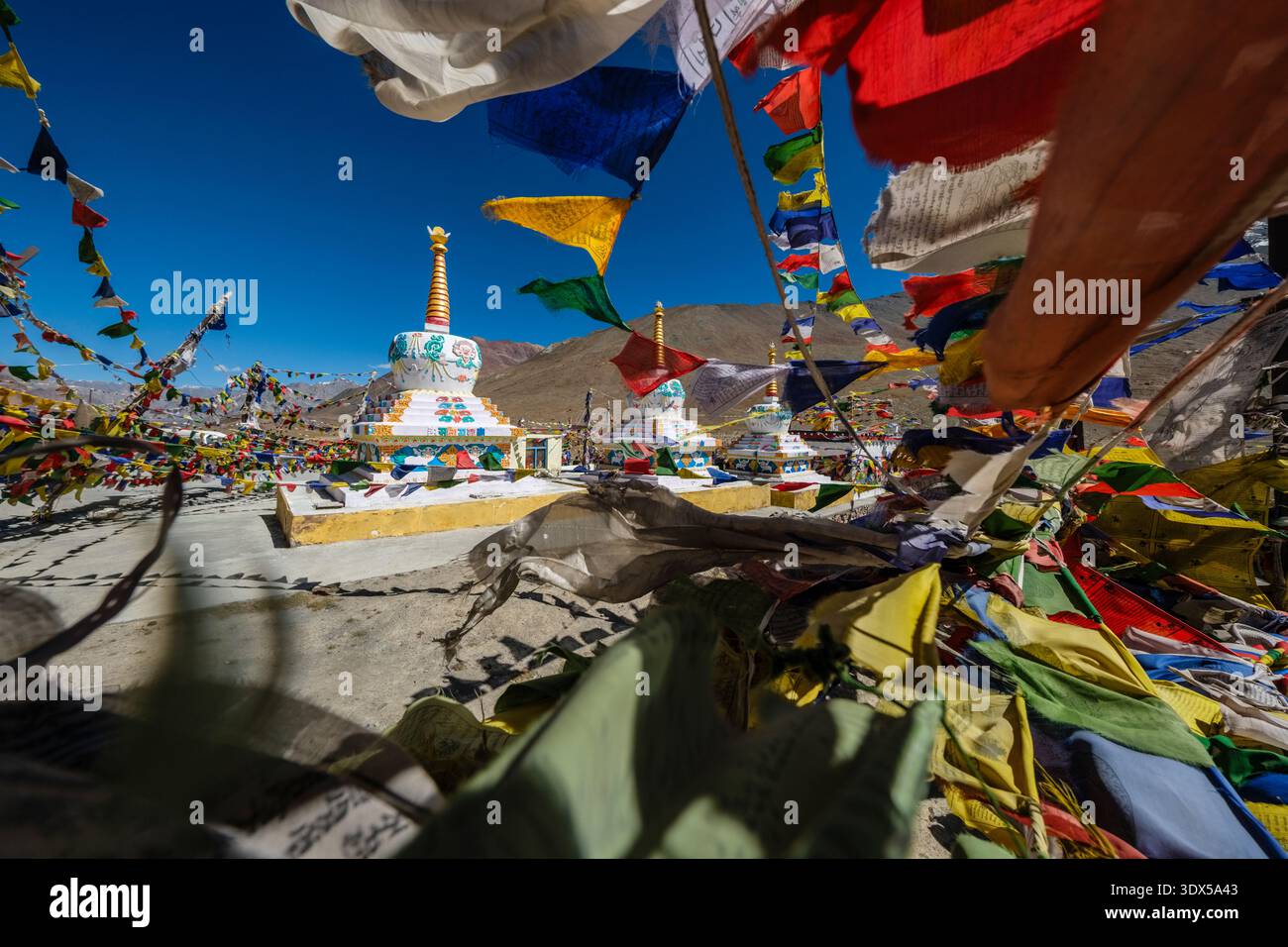 Weiße Chorten sind von bunten Gebetsfahnen umgeben, die in der Kunzum La bei Losar liegen, wobei die zerklüfteten Hänge des Himalaya in der Ferne aufsteigen. Stockfoto