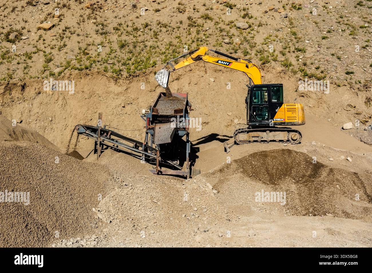 Ein SANY-Bagger lädt Kies in eine Sortiermaschine auf einer staubigen Baustelle in Sagnam, die aus verschiedenen Haufen verarbeiteter Steine besteht. Stockfoto