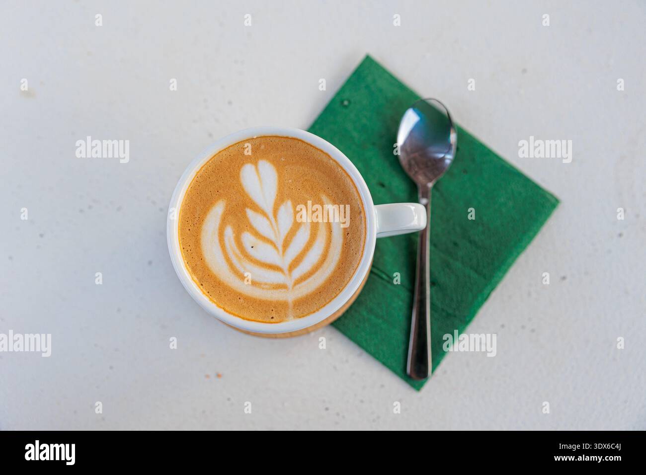 Cremiger Latte mit zarter Blattlattkunst, präsentiert in einer weißen Tasse auf einer grünen Serviette neben einem Löffel. Minimaler Overhead-Aufbau sorgt für gemütliche Morgenstunden Stockfoto