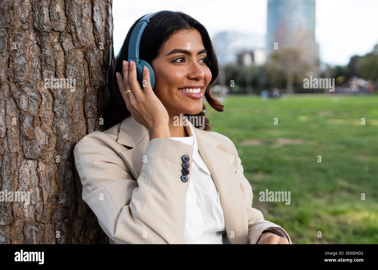 Die hispanische Frau lehnt sich an einen Baum, trägt Kopfhörer und einen beigefarbenen Blazer, lächelt und genießt Musik in einem Park mit verschwommenem Stadtbild Stockfoto