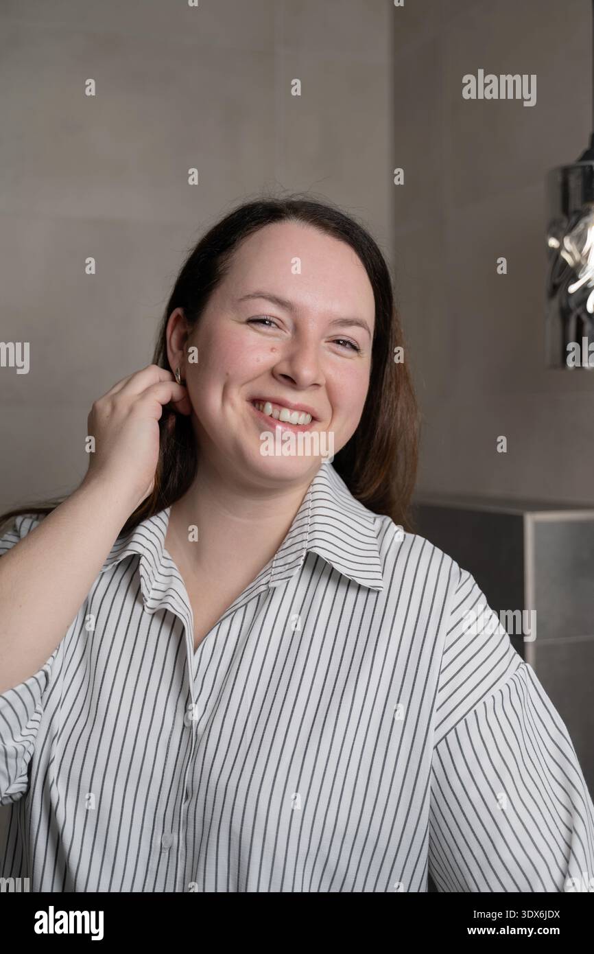 Eine fröhliche Frau mit langen Haaren lächelt in einem komfortablen Zuhause. Sie trägt ein gestreiftes Hemd und berührt beiläufig ihr Ohr, um eine Re hervorzuheben Stockfoto