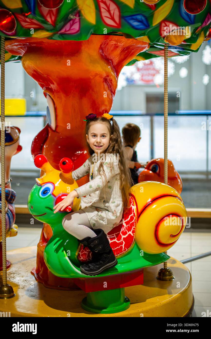 Glückliches Mädchen reitet auf farbenfrohen Karusselltieren im Einkaufszentrum Vergnügungsbereich mit hellen Lichtern und verspielter Innenatmosphäre. Stockfoto