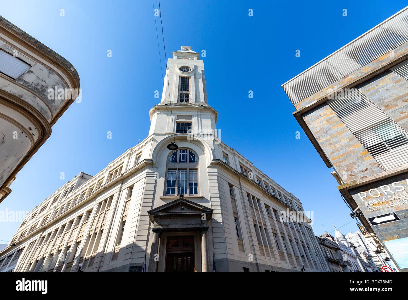Ein Niedrigwinkelbild fängt die architektonische Pracht eines hohen, weißen Gebäudes mit einem markanten Uhrenturm ein, der vor einem klaren blauen Himmel eingerahmt ist. Die Bügel Stockfoto