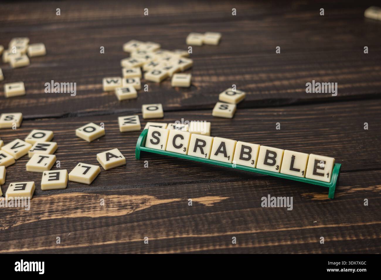 Die Fliesen sind auf einem Holztisch mit einem Scrabble-Halter verstreut, auf dem das Wort „SCRABBLE“ steht. Die Menschen genießen Spiele am National Scrabble Day zusammen. Stockfoto