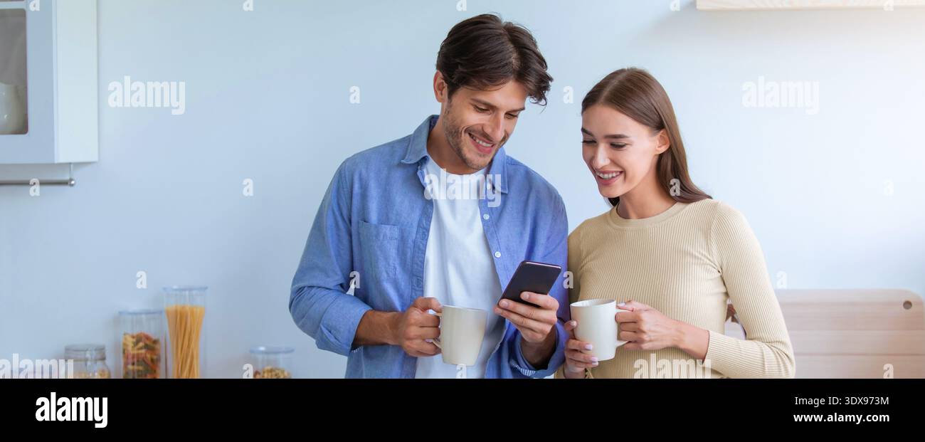 Das junge Paar teilt sich einen Moment, während es morgens in der Küche auf ein Smartphone schaut Stockfoto