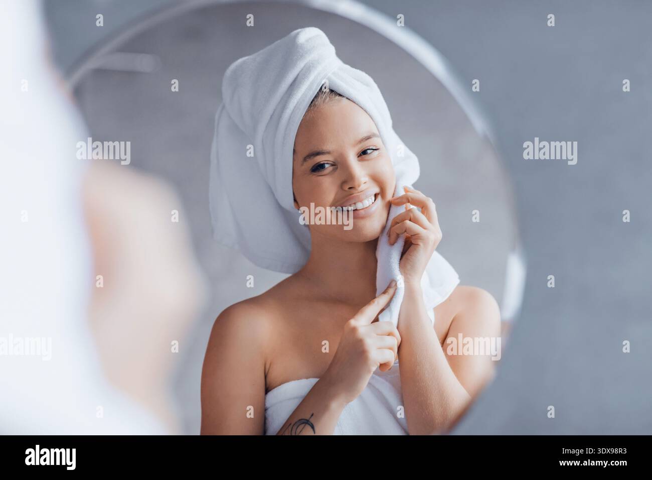 Frau, die in den Spiegel blickte, während sie lächelte, nachdem sie in einem hellen Badezimmer geduscht hatte Stockfoto