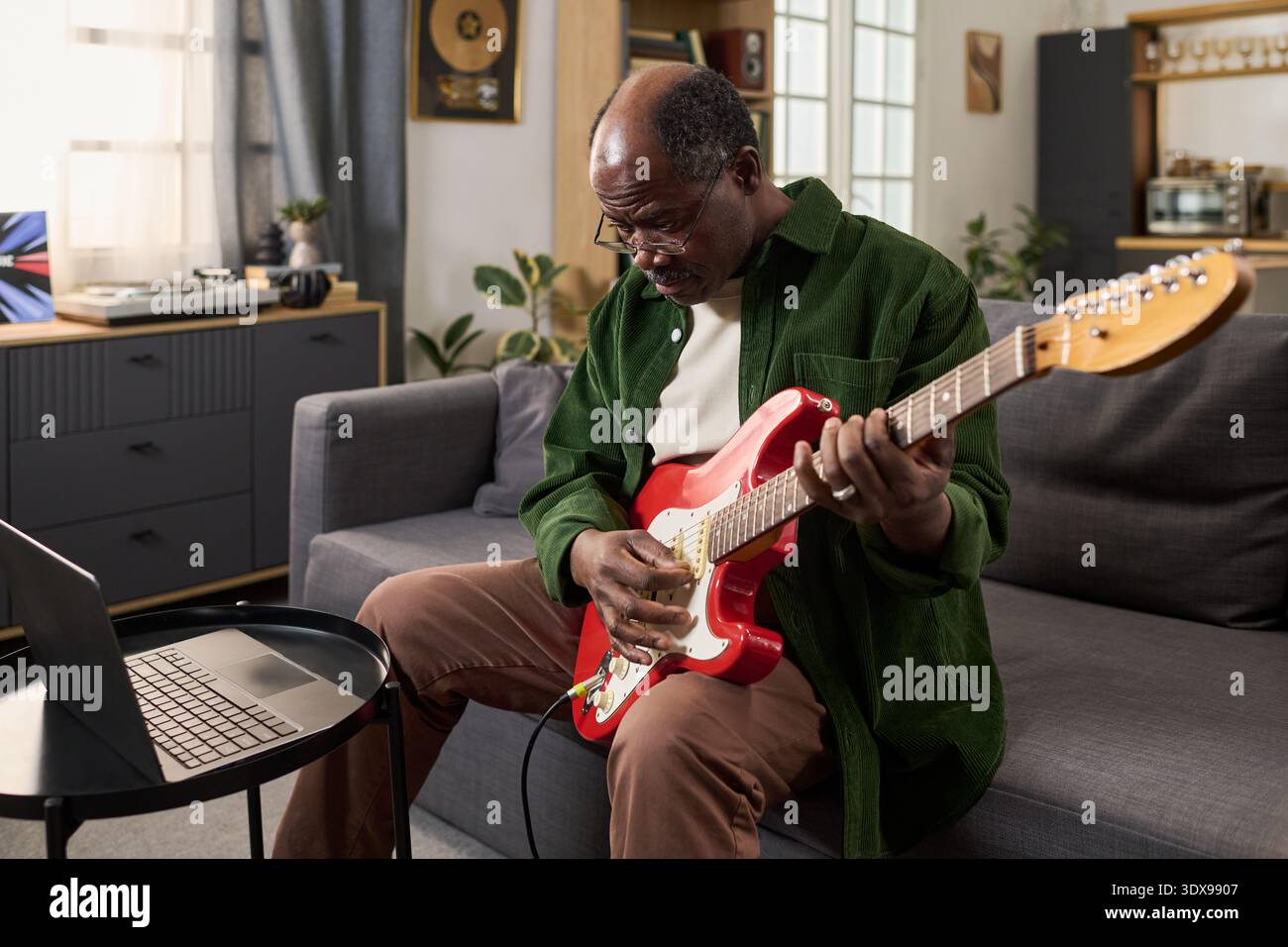 Senior Black man sitzt auf dem Sofa und spielt elektrische Gitarre, während er auf den Laptop-Bildschirm schaut, Musik zu Hause übt und sich auf das Erlernen neuer Akkorde und Techniken konzentriert Stockfoto
