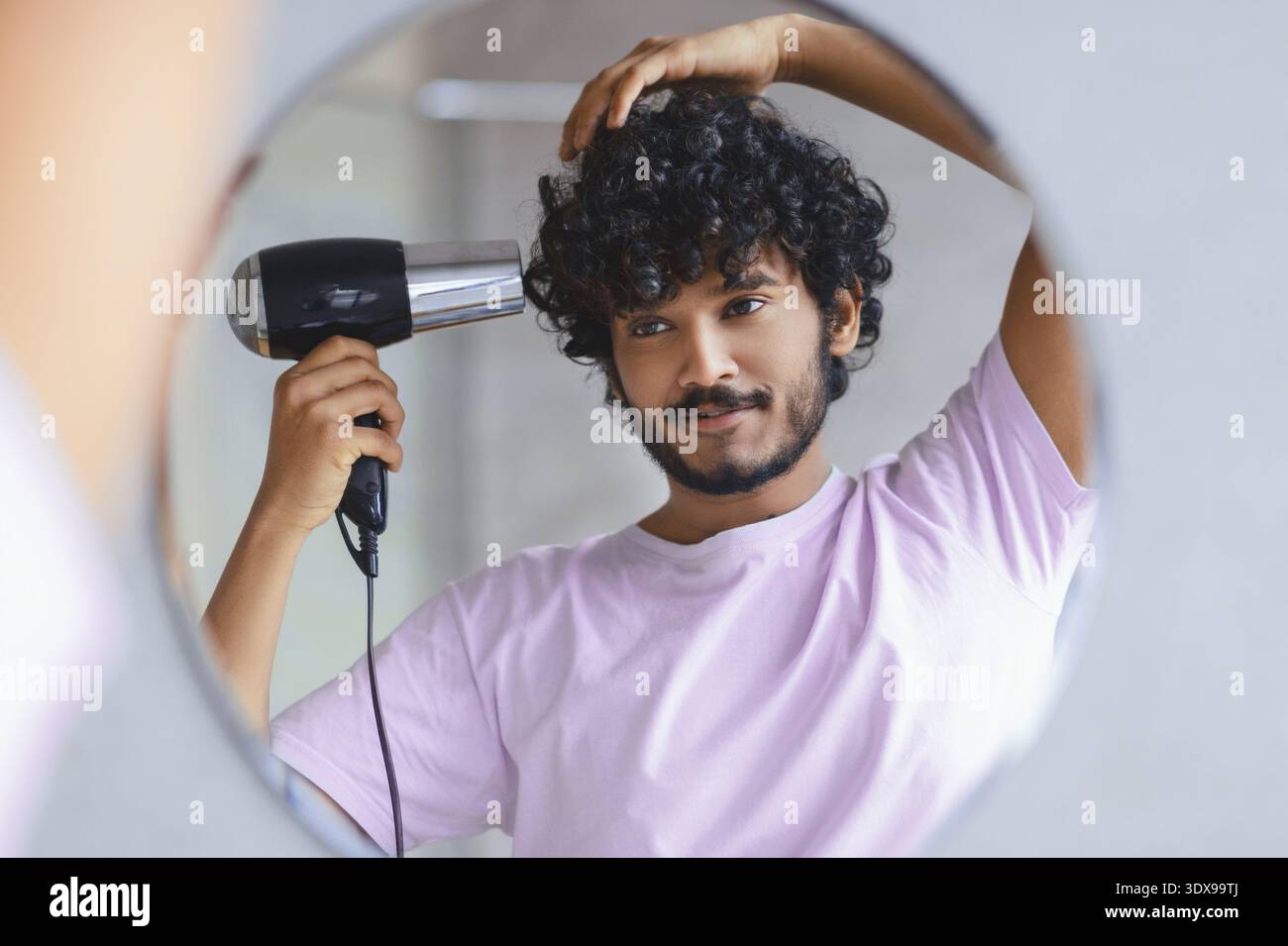 Junger Mann stylt sein lockiges Haar vor einem Spiegel mit einem Haartrockner Stockfoto