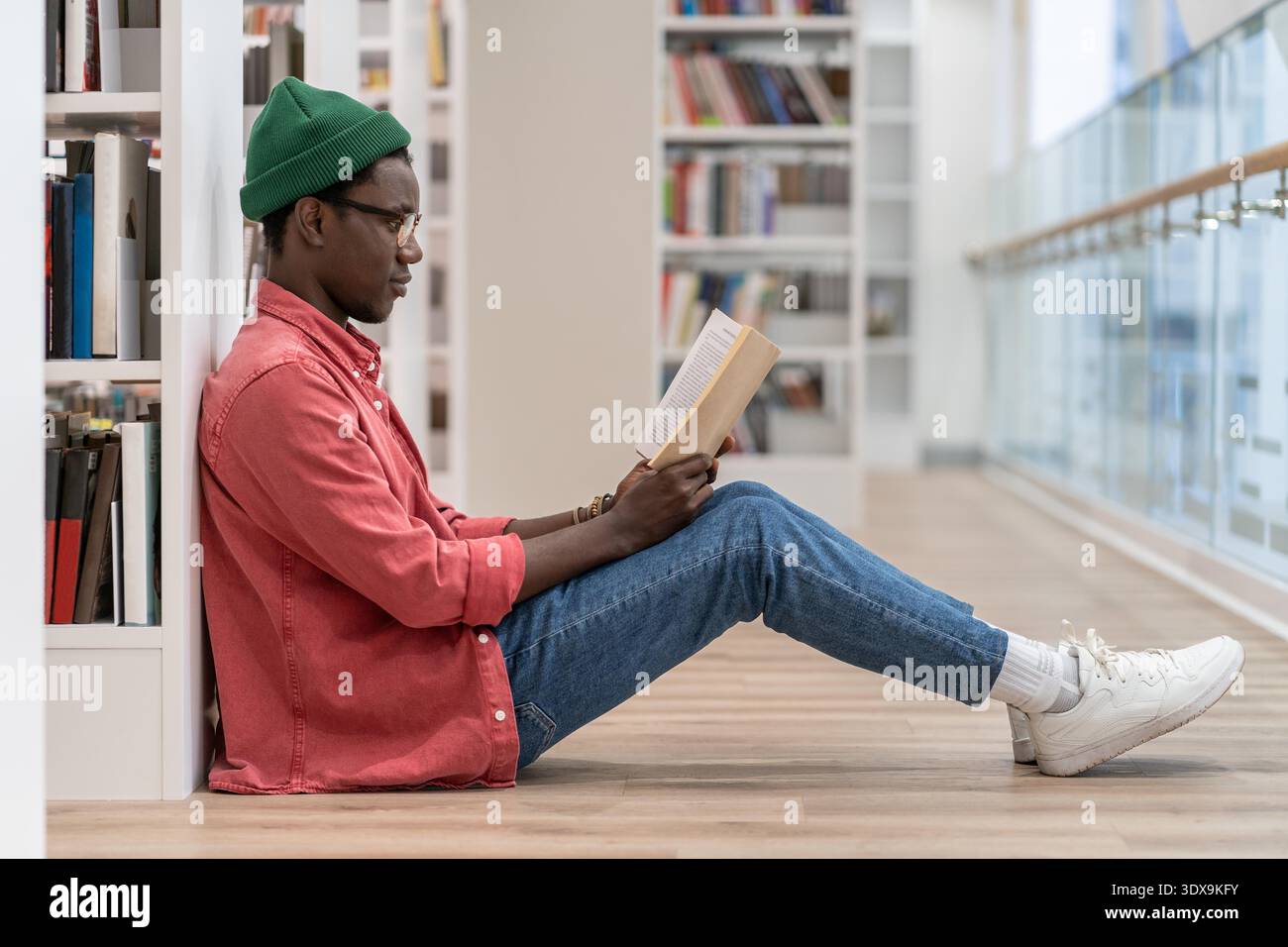 Der gebündelte Schwarze Studentin, der Buchliebhaber, der freie Freizeit in der Bibliothek verbringt, sitzt Afroamerikaner mit Buch in der Hand auf dem Boden in der Nähe des Bücherregals und genießt das Lesen. Hobby, Bildung, Universitätskonzept || Modell veröffentlicht Stockfoto