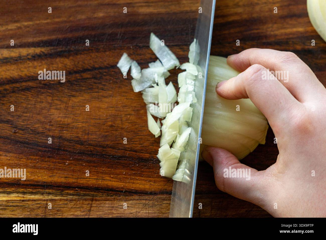 Nahaufnahme einer Hand, die weiße Zwiebeln mit einem Küchenmesser auf einem hölzernen Schneidebrett hackt. Stockfoto