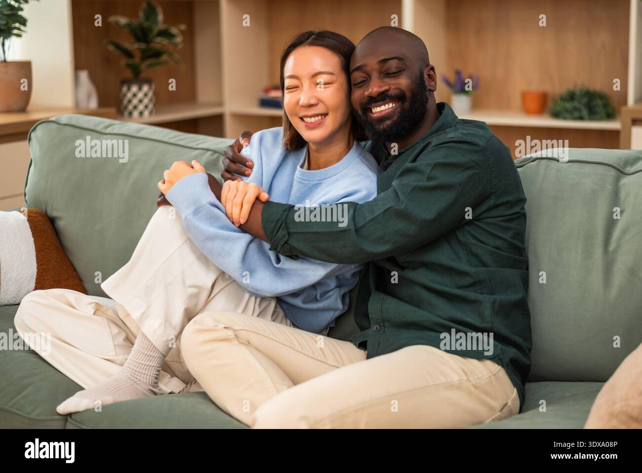 Ein fröhliches, multirassisches Paar sitzt eng zusammen auf einem grünen Sofa, lacht und umarmt in einem gemütlichen, modernen Wohnzimmer. Stockfoto