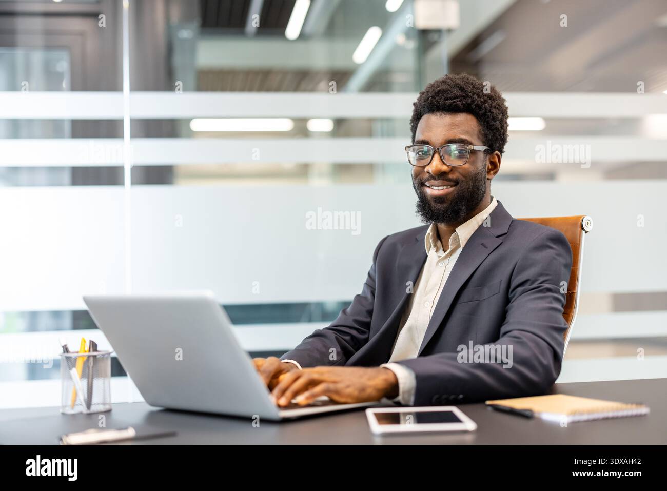Junger professioneller Mann mit Brille, der auf einem Laptop schreibt, in die Kamera blickt und lächelt, während er an seinem Schreibtisch in einer modernen Unternehmensumgebung sitzt Stockfoto