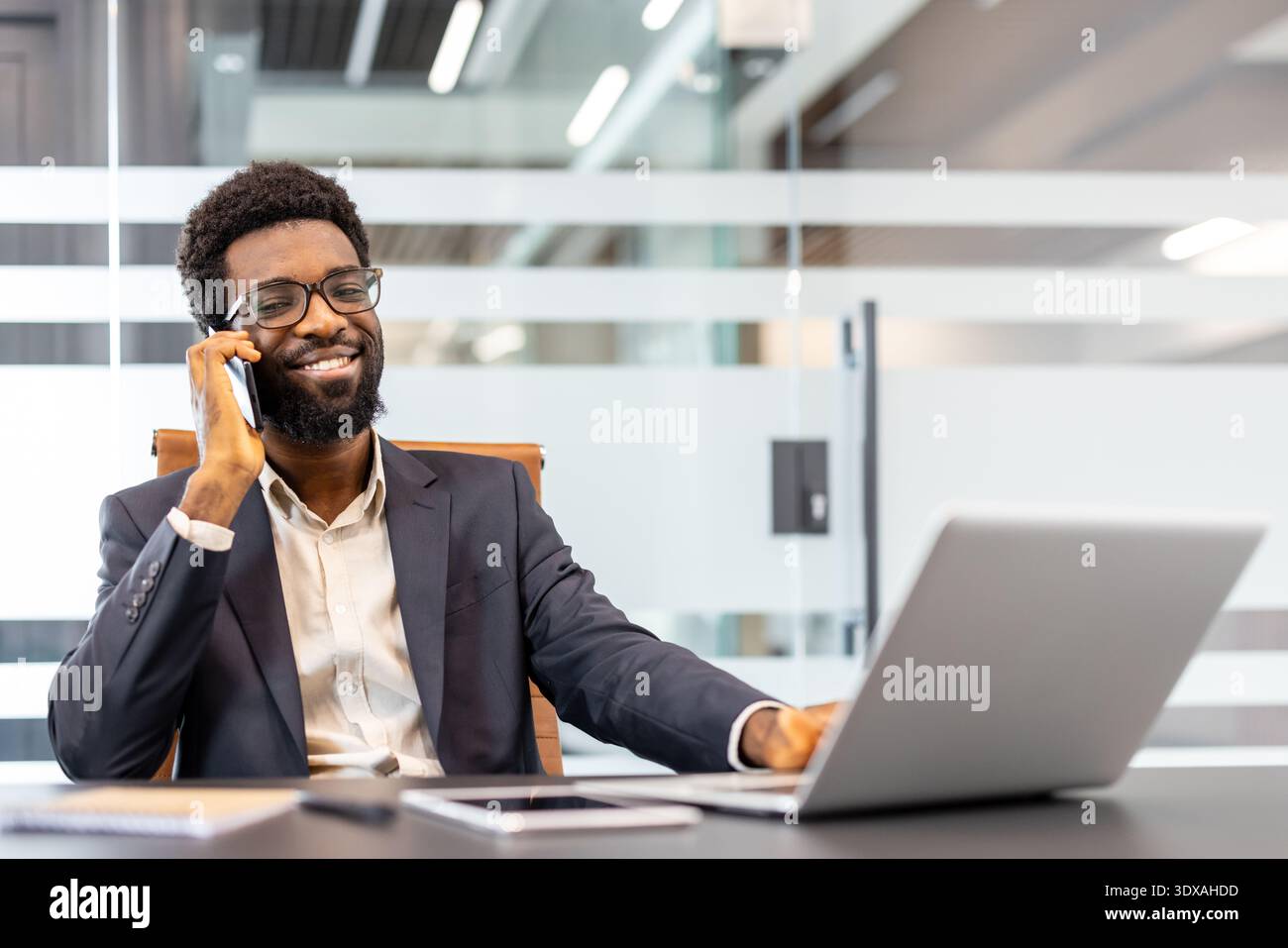Lächelnder professioneller Mann, der am Handy spricht, während er in einem modernen Büro an einem Laptop arbeitet, sich für eine klare Kommunikation und erfolgreiche Geschäftsbeziehungen einsetzt Stockfoto
