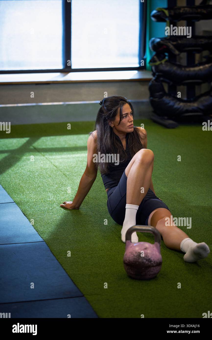 Müde Frau, die wegschaut, während sie im Fitnessstudio auf einer Grasmatte sitzt Stockfoto