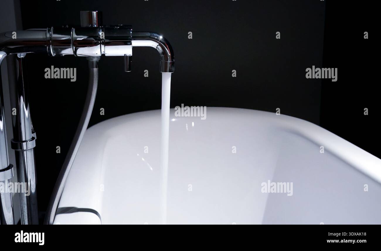 Nahaufnahme eines modernen Chrom-Wasserhahns mit einem konstanten Strom von frischem Wasser, das in eine weiße Keramikbadewanne fließt. Das Bild zeigt ein minimalistisches DE Stockfoto