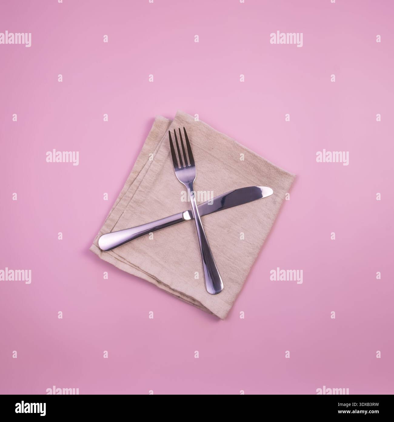Eine Gabel und ein Messer werden auf einer beigen Serviette auf einem rosafarbenen Hintergrund platziert. Die Utensilien sind aus Edelstahl mit glänzender Oberfläche. Stockfoto