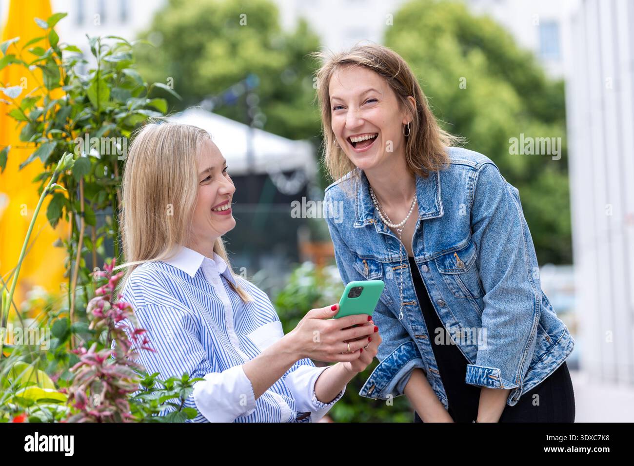 Zwei Frauen lachen zusammen auf sonniger Terrasse Grün Pflanzen Blumen teilen Smartphone lebhafte Gespräche. Legere Outfits warmes Tageslicht fröhlich Stockfoto