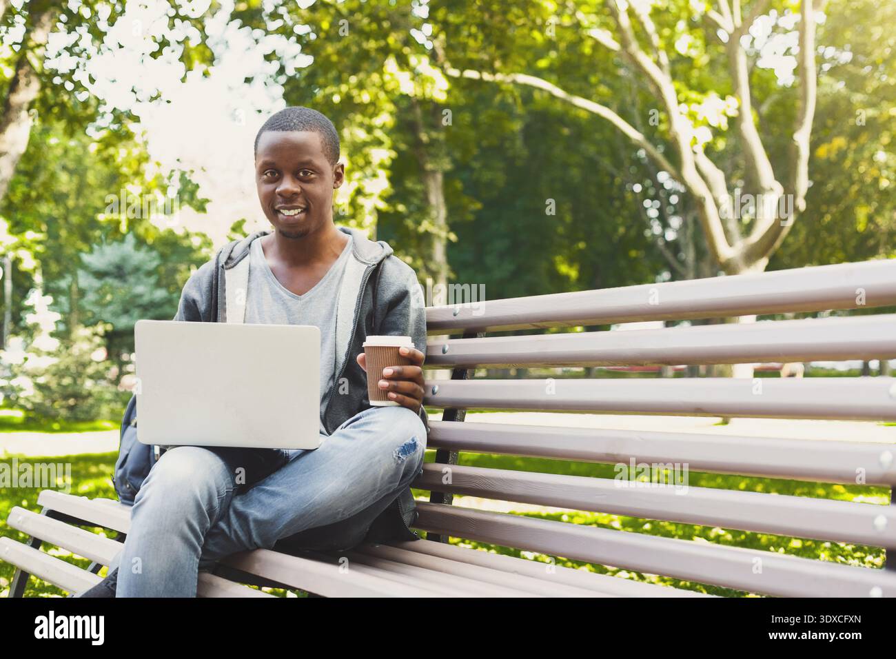 Junger Mann, der an einem Laptop im Park arbeitet, während er an einem sonnigen Tag eine Kaffeetasse hält Stockfoto
