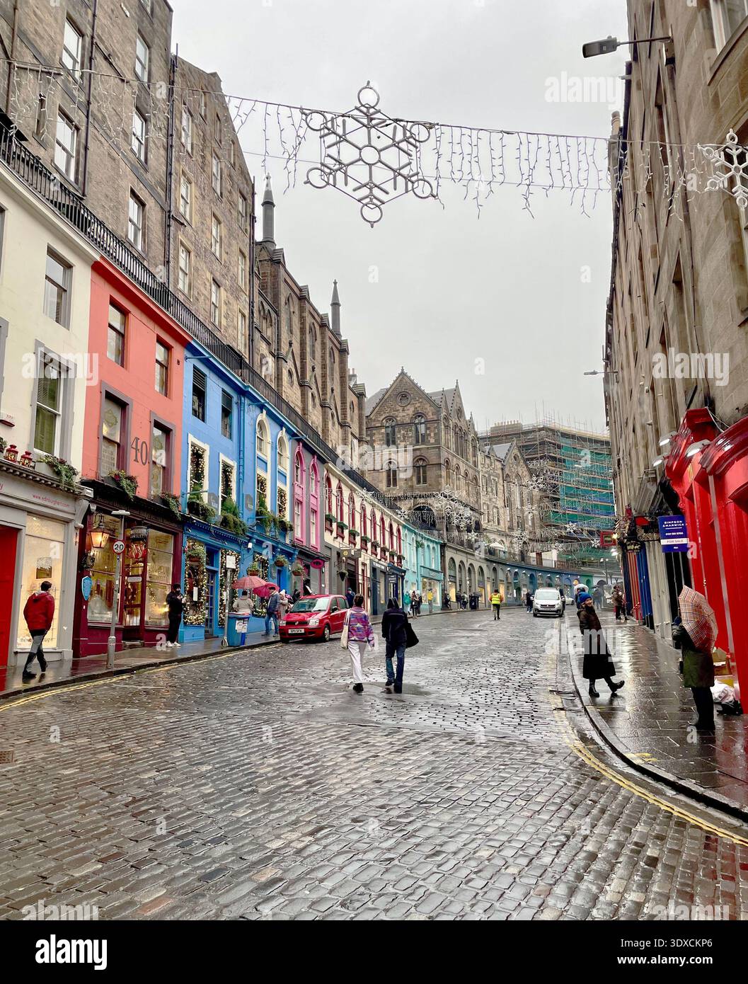 Fußgänger spazieren entlang der Victoria Street in Edinburghs Altstadt, einer farbenfrohen kopfsteingepflasterten Straße, die von Geschäften und festlichen Lichtern gesäumt ist. - Smartphone-aufgenommenes Stockfoto