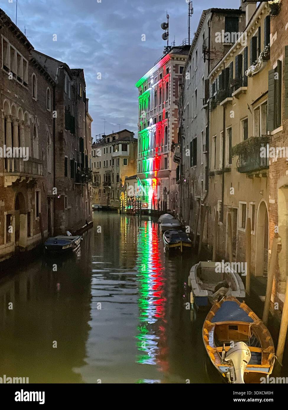 Italienische Flaggenfarben beleuchten ein Gebäude entlang eines ruhigen venezianischen Kanals in der Abenddämmerung und spiegeln sich auf dem Wasser neben verankerten Booten wider. - Smartphone-aufgenommenes Stockfoto