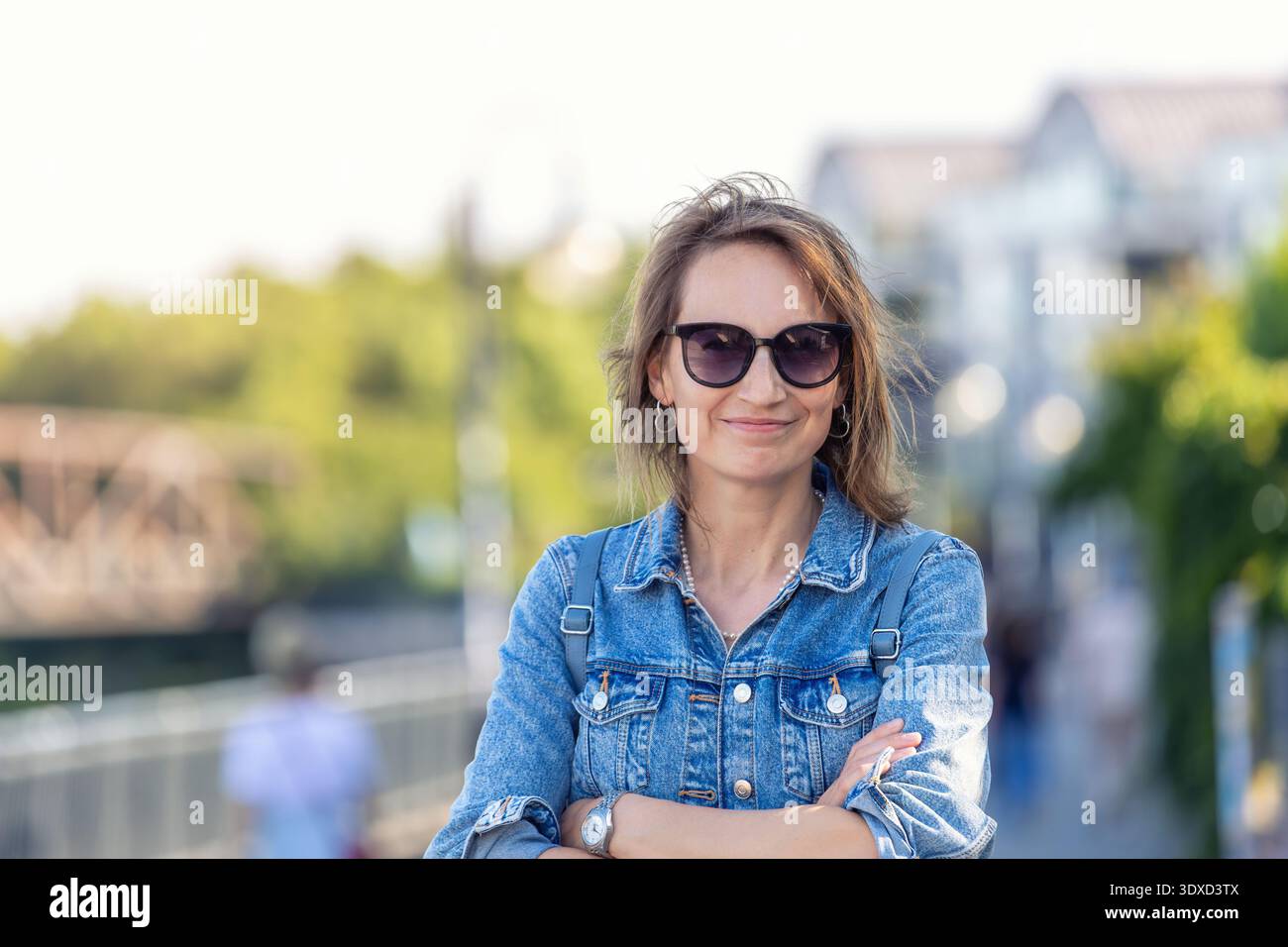 Selbstbewusste Frau tragen Sonnenbrille Denim-Jacke steht in der Stadtstraße nahe der Flussbrücke und drückt Unabhängigkeit und modernen Lebensstil aus. Outdoor Porträt Sommer Stockfoto
