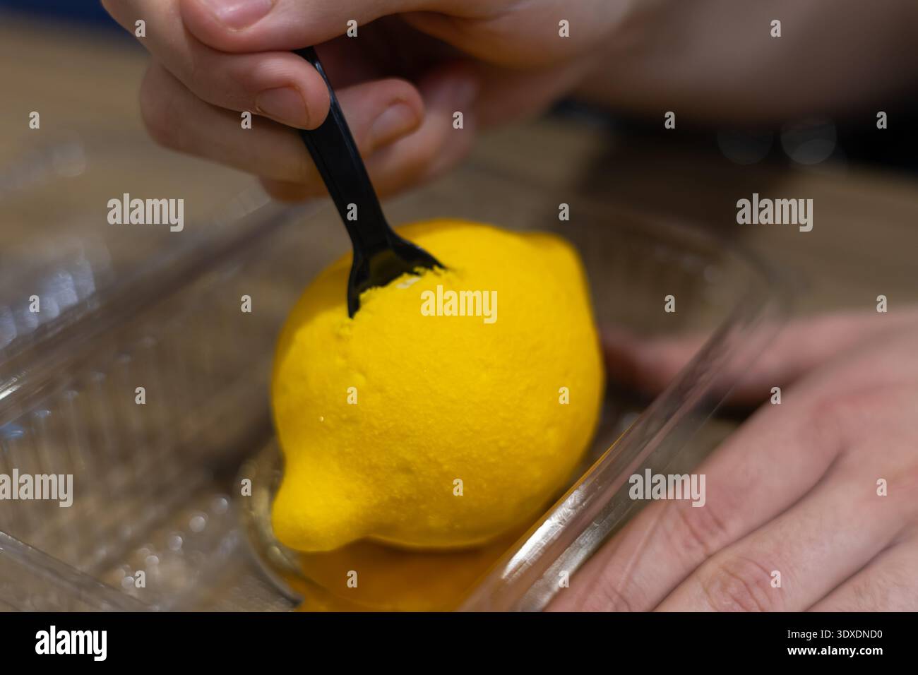 Person, die eine schwarze Gabel verwendet, um ein realistisches Zitronenmousse-Dessert zu probieren, das in einem Plastikbehälter serviert wird, Nahaufnahme von lebendigem gelbem Zitrusgebäck Stockfoto
