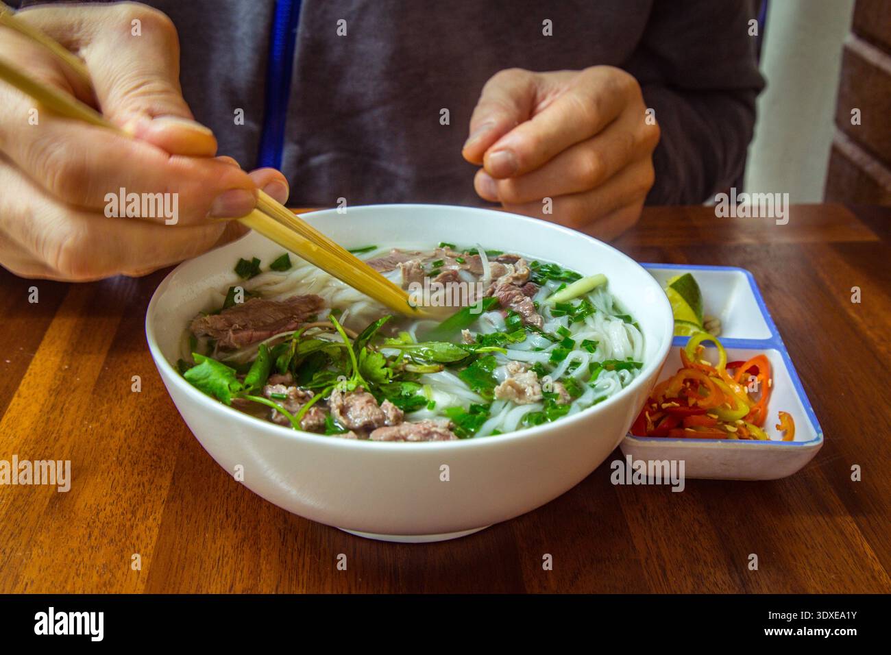 Ein Mann isst traditionelle vietnamesische Rindersuppe Pho Bo mit Essstäbchen. Street Food. Eine Schüssel Pho mit Nudeln und Rindfleisch essen. Mann hält Essstäbchen Stockfoto