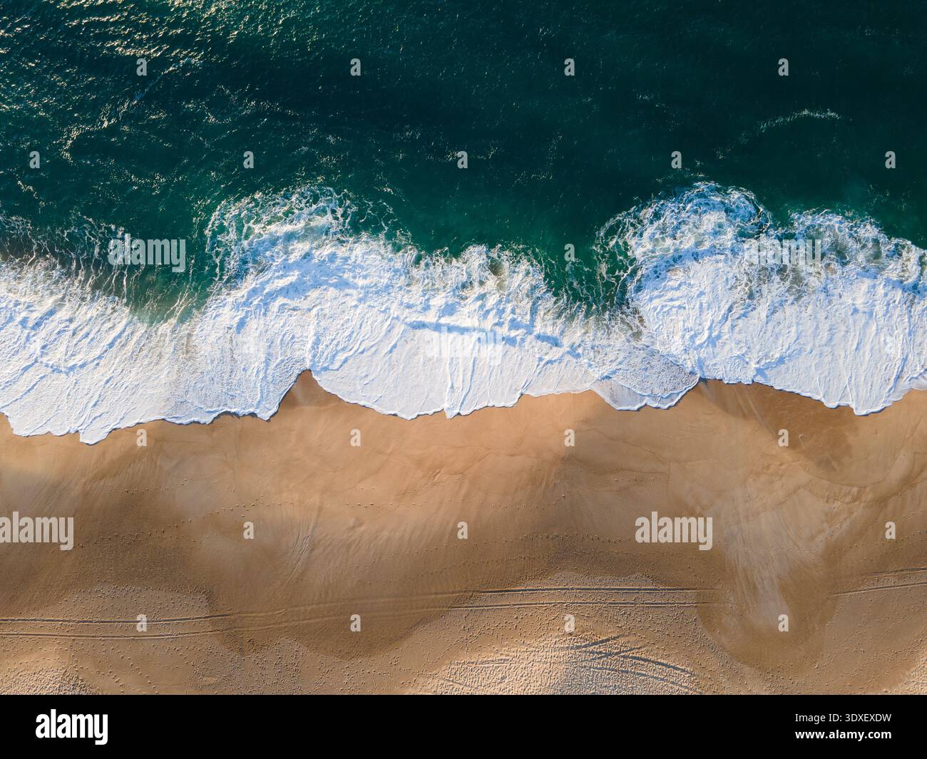 Aus der Vogelperspektive sehen Sie die pulsierenden türkisfarbenen Wellen, die auf den goldenen Sand stürzen und einen faszinierenden Kontrast von Farbe und Textur schaffen, Nazaré, Leiria Distri Stockfoto