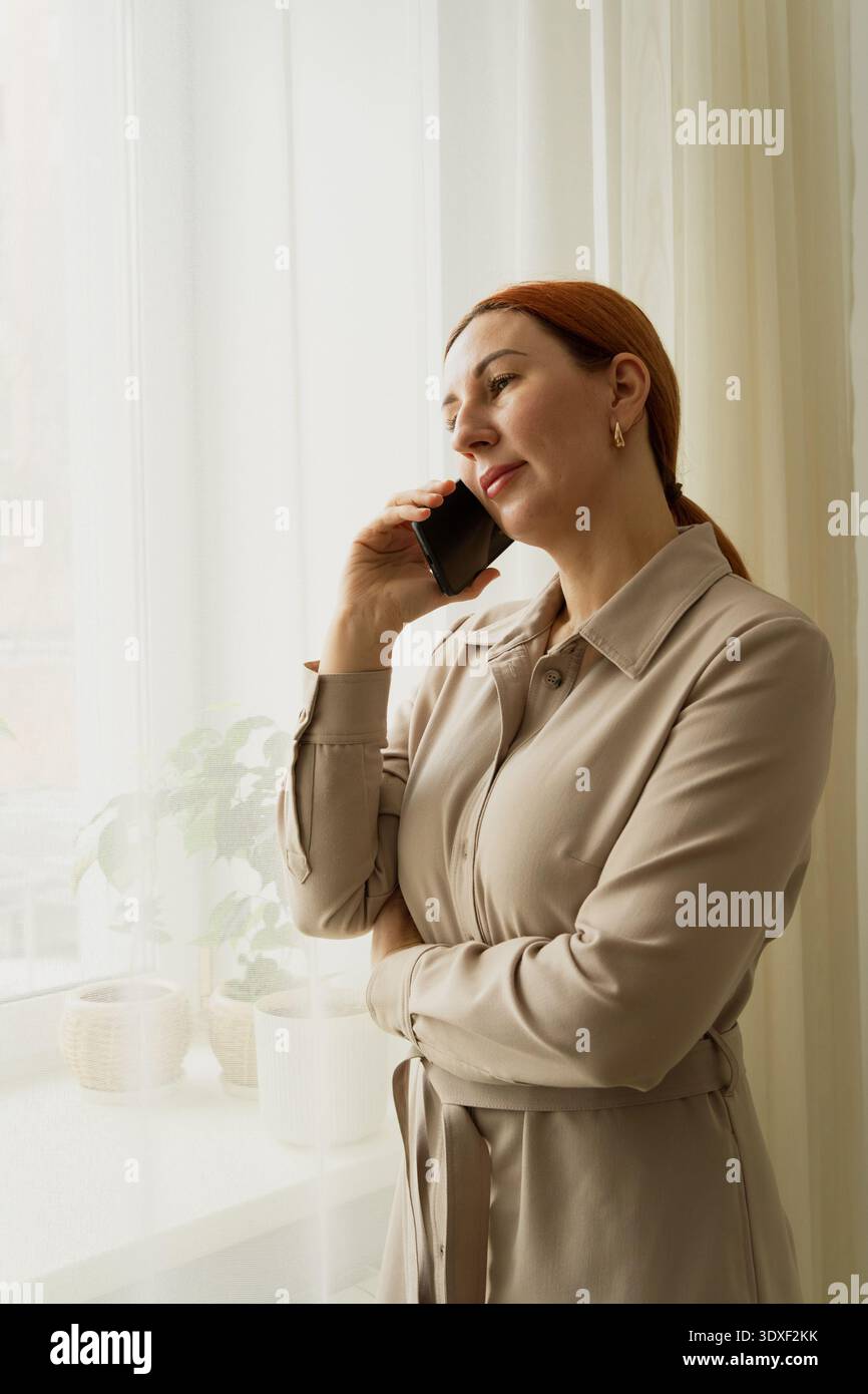 Frau führt ein Geschäftsgespräch, steht mit ihrem Smartphone am Fenster, fühlt sich nachdenklich und kontaktfreudig, während sie von zu Hause aus arbeitet Stockfoto