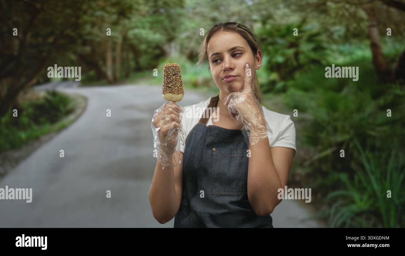 Frau mit Eis und Schürze mit Einweghandschuhen, Finger an Wange auf einer Waldstraße; Versuchungszweifel. Stockfoto