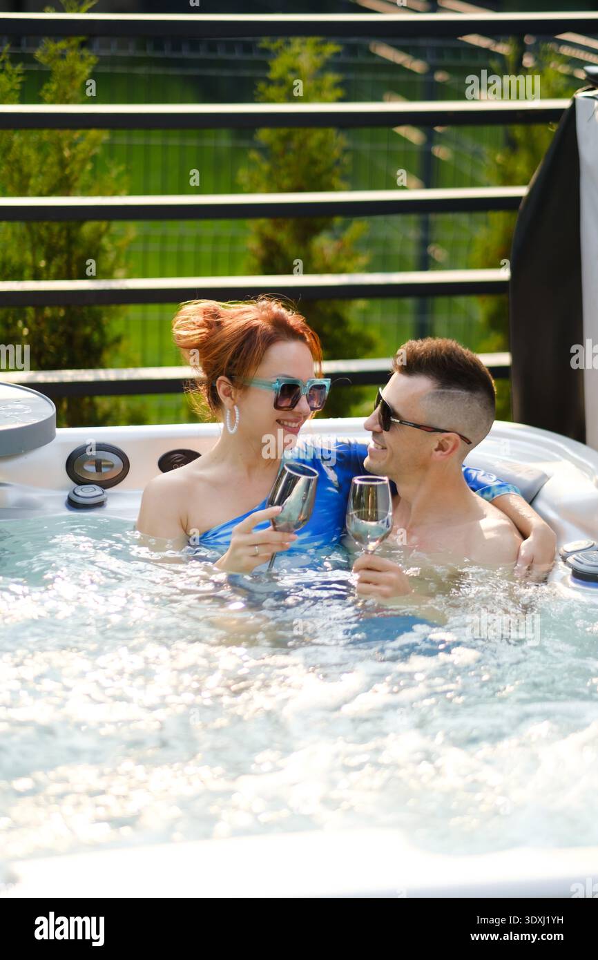 Zwei Personen entspannen sich in einem Whirlpool. Sie halten Drinks und lächeln einander an. Es ist ein sonniger Tag. Die Szene zeigt einen Balkonblick mit Grün im Backgr Stockfoto