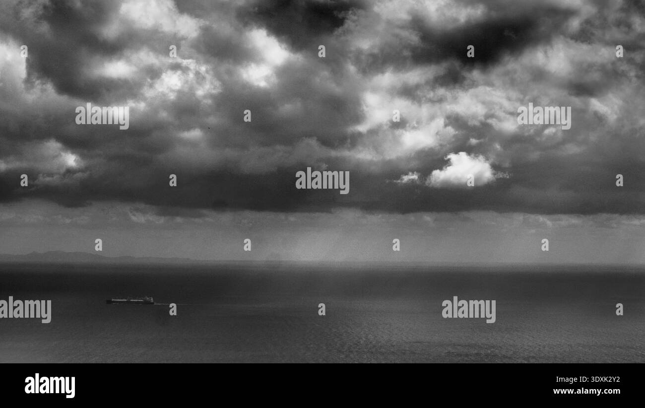 Einfarbige Meereslandschaft mit einem einsamen Frachtschiff, das ruhiges Wasser unter schweren Sturmwolken mit sichtbaren Lichtstrahlen am Horizont überquert. Konzeptbild Stockfoto