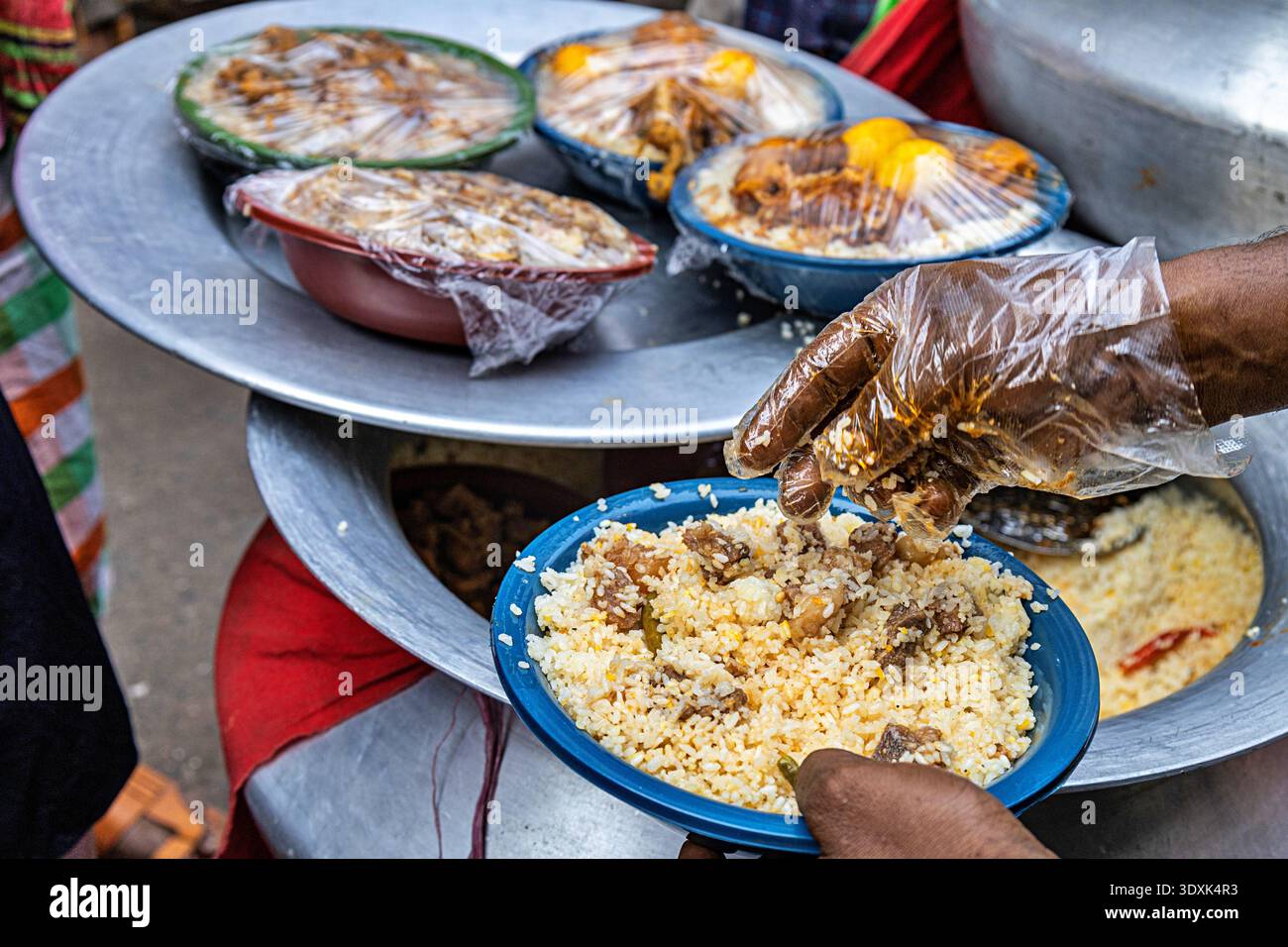 Blick auf Dampfplatten und Schüsseln, die hoch mit Reis und Fleisch gefüllt sind, ein Verkäufer, der Speisen im belebten Chawkbazar, Dhaka, Dhaka Division, Bangladesch serviert. Stockfoto