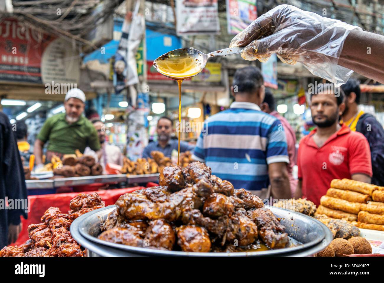 Blick auf den goldenen Sirup, der von einem Löffel auf einen Haufen glitzernder, dunkel glasierter Leckereien inmitten des geschäftigen Chawkbazar Markts in Dhaka, Bangladesch. Stockfoto