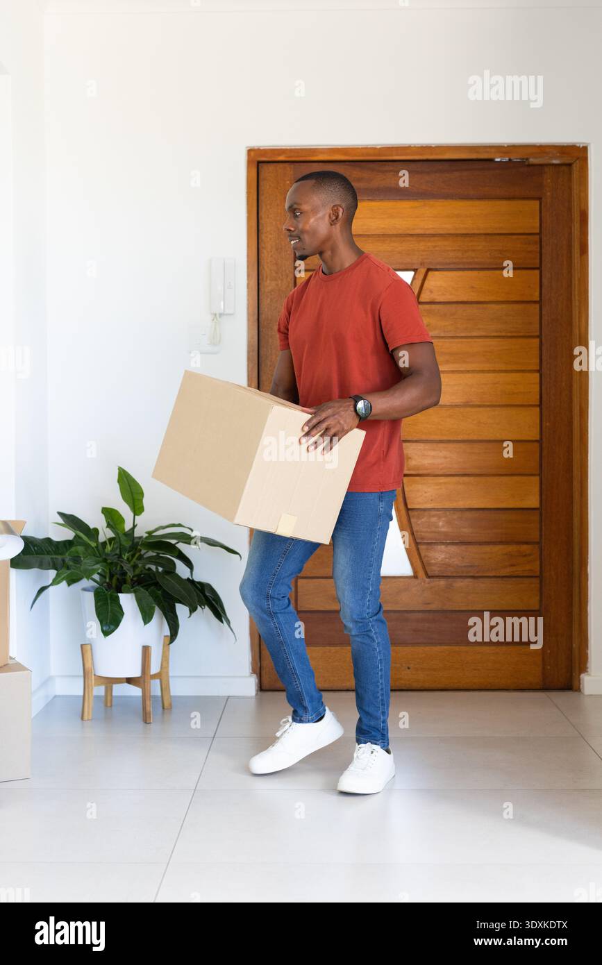 Erwachsener Mann mit rotem T-Shirt und Jeans mit Pappkarton, während er im Eingangsbereich neben der Pflanze steht Stockfoto