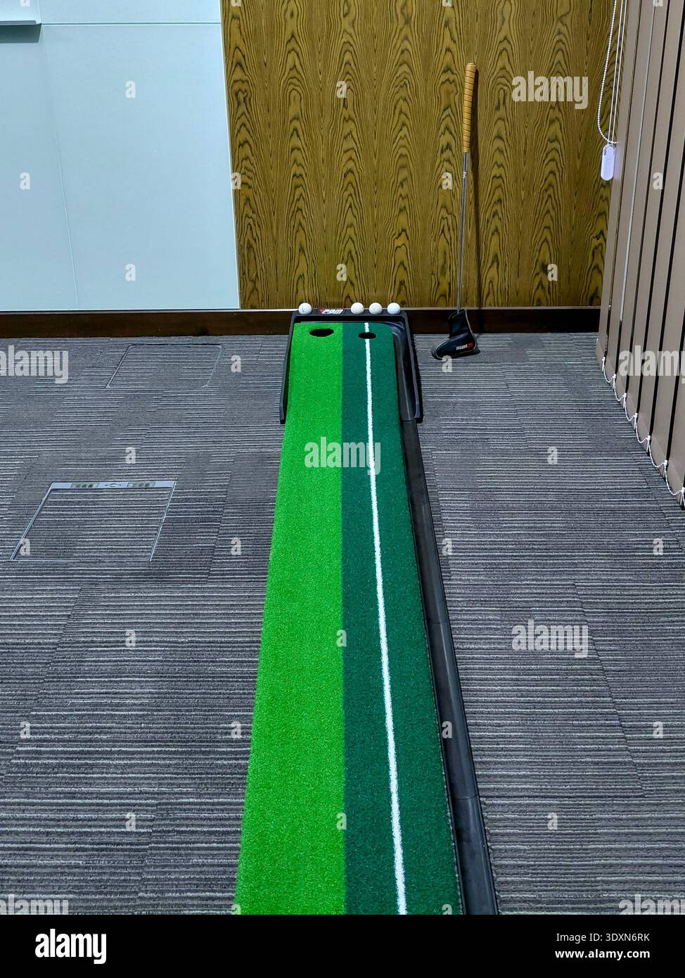 Indoor Golf Putting Übungs-Set mit PGM Green Matte, weißen Golfbälle und Putter, der sich gegen Holzmaserwand in Büro- oder Heimtrainingsumgebung lehnt Stockfoto