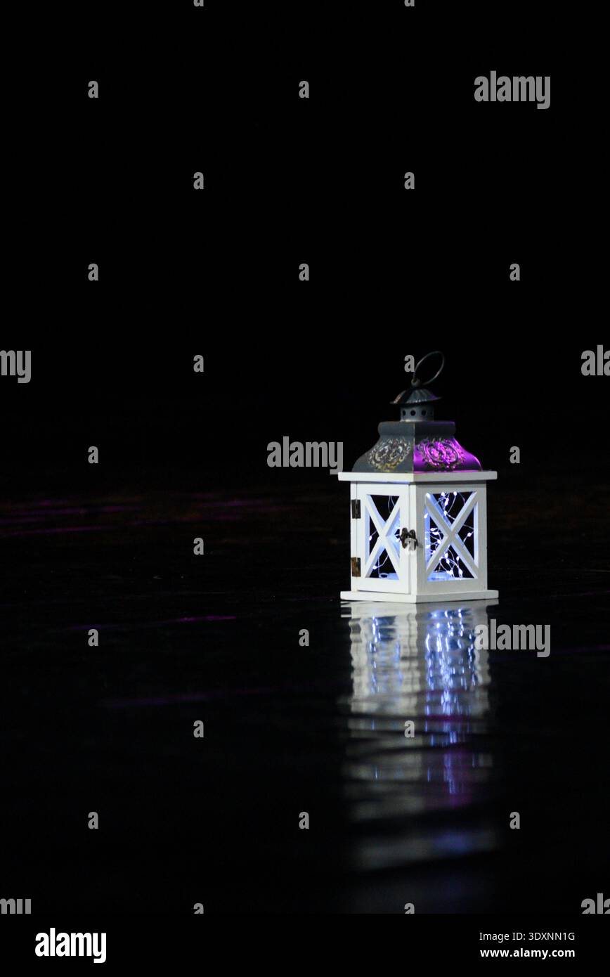 Elegante weiße dekorative Laterne, beleuchtet mit sanftem lila Licht, stehend auf einer reflektierenden dunklen Oberfläche. Minimale, stimmungsvolle Komposition mit Kopierraum Stockfoto