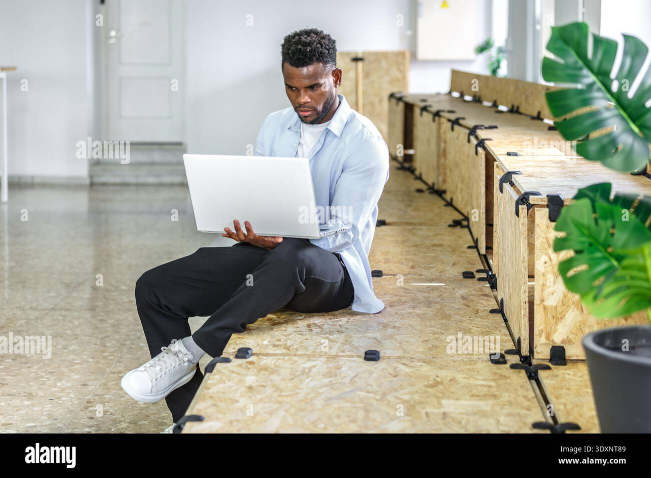 Ein junger Mann sitzt auf einer Holzbank und konzentriert sich auf seinen Laptop in einem hellen und luftigen Coworking-Raum. Tageslicht erfüllt den Raum, während Innenpflanzen eine zusätzliche Note verleihen Stockfoto